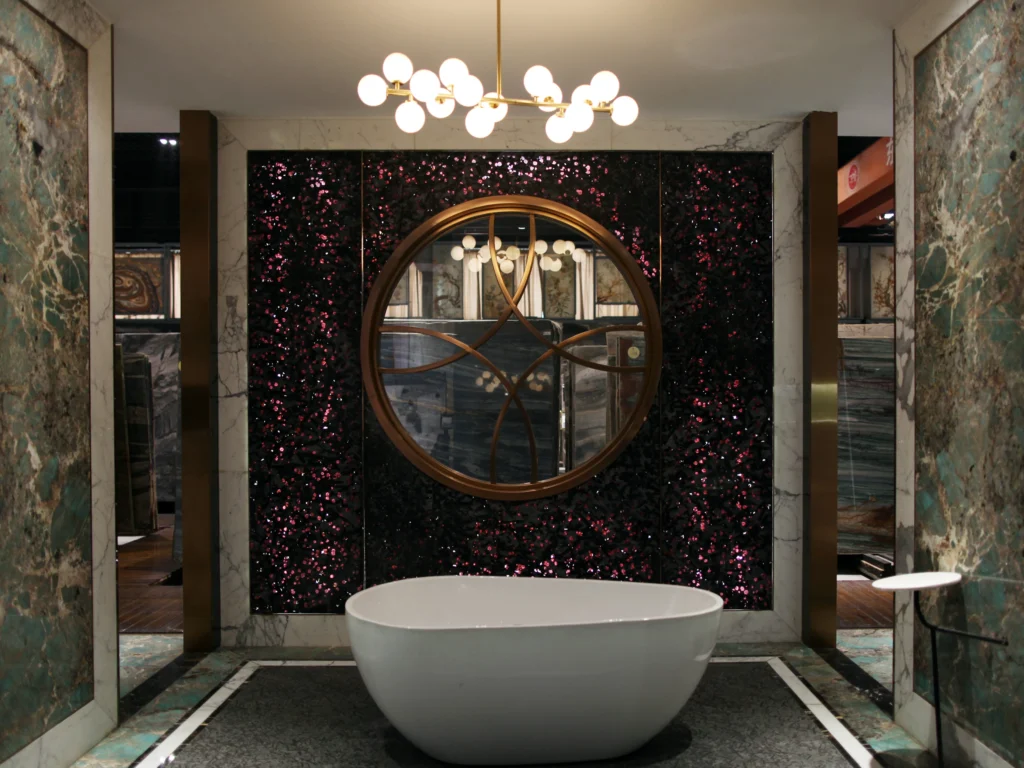 Dongxing Luxury Stone Bathroom Display: Freestanding white tub, glittering pink-flecked stone wall, and gold-framed round mirror for a lavish aesthetic.